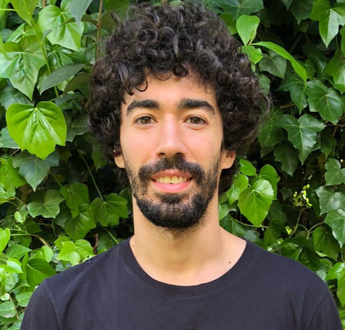 Portrait photo of a man with curly dark hair standing in front of greenery.