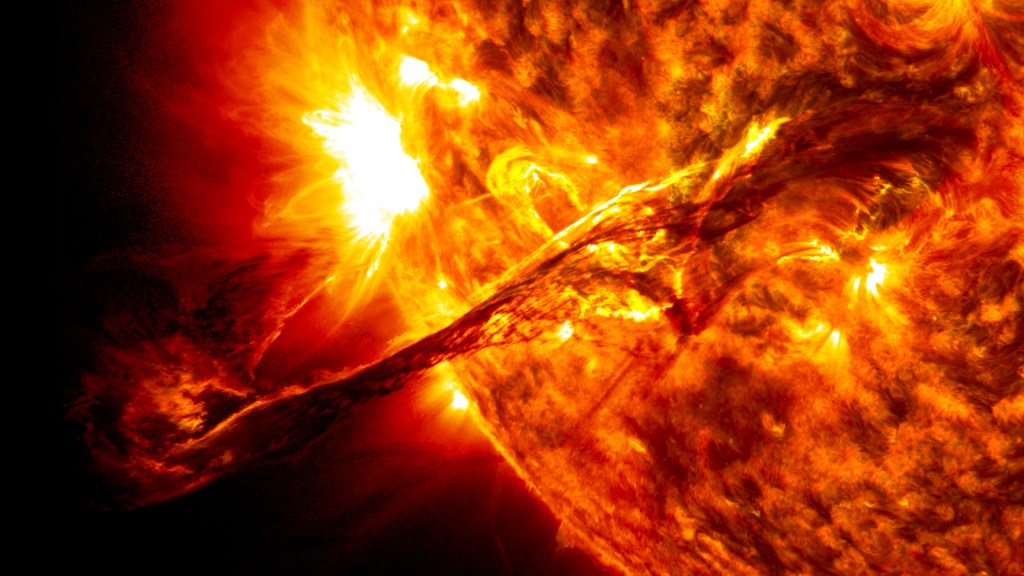 Seen against a black background, a long filament extends off the Sun. The Sun is in reds and yellows. The filament is thing and extends off the Sun, against the black background.